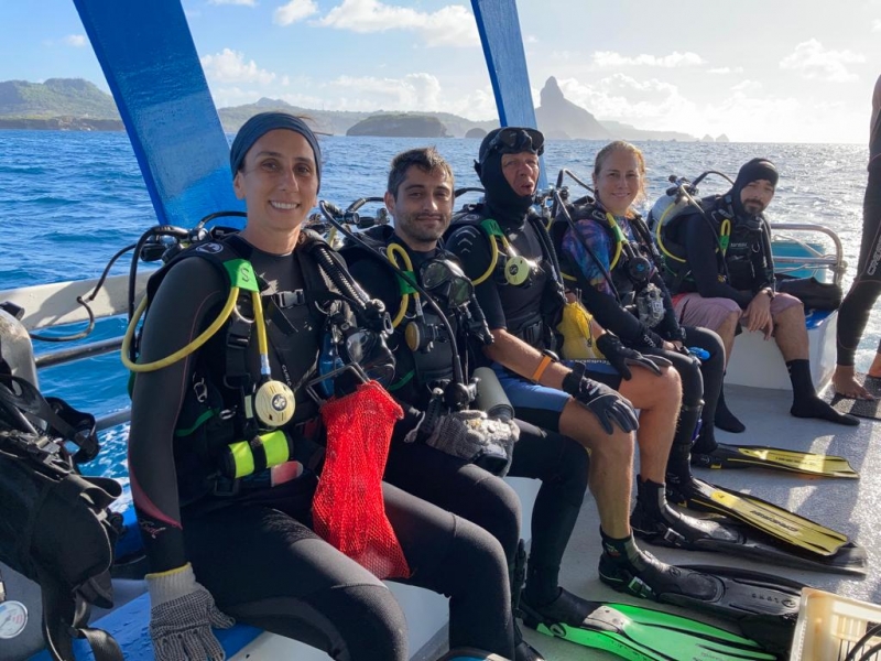 Cinco pessoas vestidas com equipamento de mergulho sentadas em um barco, olhando para a foto
