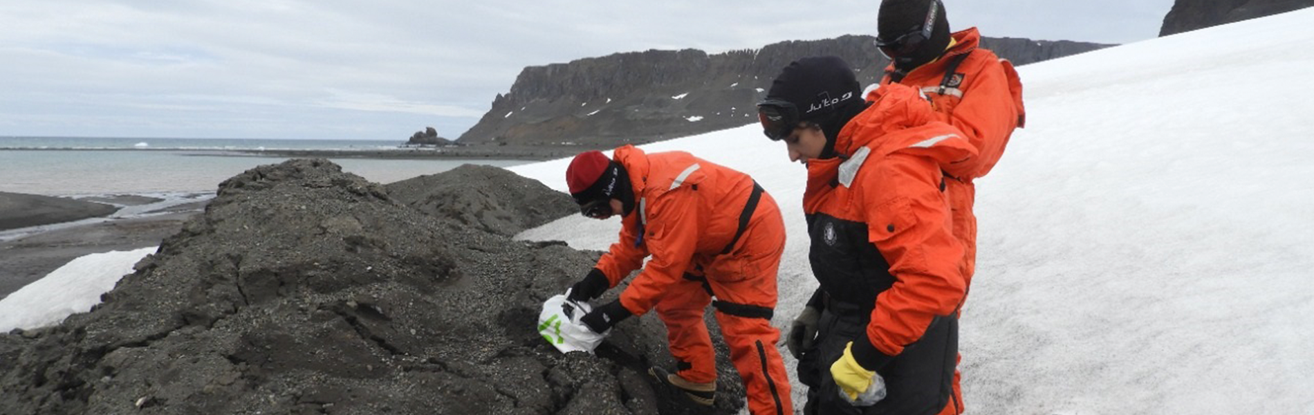 Foto de três pesquisadores vestidos de laranja examinando uma rocha em meio a neve.