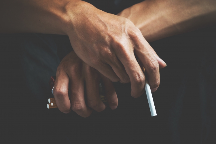 Foto de duas mãos cruzadas, uma segurando um cigarro e outra segurando um maço de cigarros