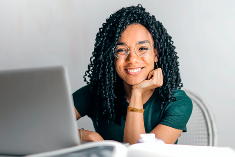 Mulher negra de óculos sorrindo sentada em frente a mesa com laptop