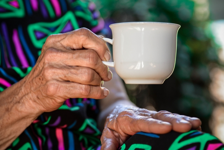 Mão direita de uma mulher idosa segurando uma xícara branca. Ao fundo sua outra mão está apoiada sobre a perna, recoberta por um tecido estampado.