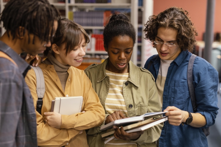 Foto de 4 estudantes universitários negros e brancos, em pé, fazendo a leitura de um livro