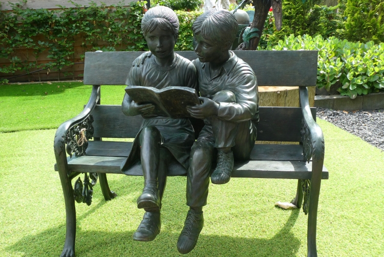Escultura de duas crianças lendo um livro em uma cadeira, dentro de um jardim