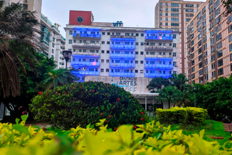 Prédio da reitoria da UFF iluminado com luzes azuis. Diante dele, um jardim com plantas em diferentes tons de verde.