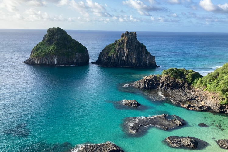 Arquipélogo de Fernando de Noronha. Na imagem se vê algumas ilhas rochosas envoltas por um mar azul esverdeado.