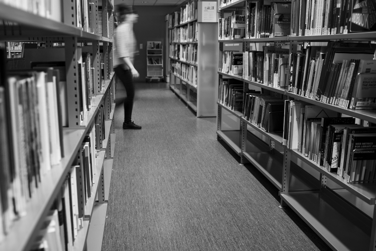 foto de corredor de biblioteca com estantes e livros, homem passando ao fundo