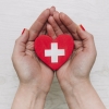 imagem: par de mãos segurando um coração vermelho com o desenho de uma cruz branca