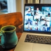 Foto de laptop aberto em cima de uma mesa com a tela mostrando um programa aberto de webconferência. Ao lado do laptop há uma caneca verde.