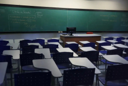 #ParaTodosVerem A imagem mostra uma sala de aula, com carteiras azuis e um quadro verde ao fundo onde podem ser vistas anotações feitas a giz