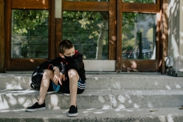 Criança de máscara sentada em um degrau, em frente a uma porta de vidro fechada.