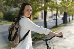 Mulher de cor branca e cabelo preto, com mochila nas costas e blusa branca, segurando sua bicicleta