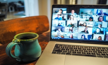 Foto de laptop aberto em cima de uma mesa com a tela mostrando um programa aberto de webconferência. Ao lado do laptop há uma caneca verde.