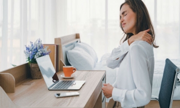 Mulher sentada em escrivaninha de frente para um laptop com a mão direita massageando o ombro esquerdo e expressão facial de desconforto