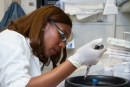 Mulher negra com roupa e luva branca, de perfil, manipulando um instrumento num laboratório