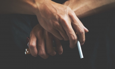 Foto de duas mãos cruzadas, uma segurando um cigarro e outra segurando um maço de cigarros