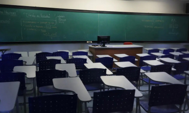 #ParaTodosVerem A imagem mostra uma sala de aula, com carteiras azuis e um quadro verde ao fundo onde podem ser vistas anotações feitas a giz