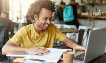 Estudante universitário negro sorrindo e sentado na frente de um computador com livros na mesaros