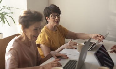 duas mulheres brancas, de terceira idade, sentadas a mesa, cada uma com seu computador e copo de café