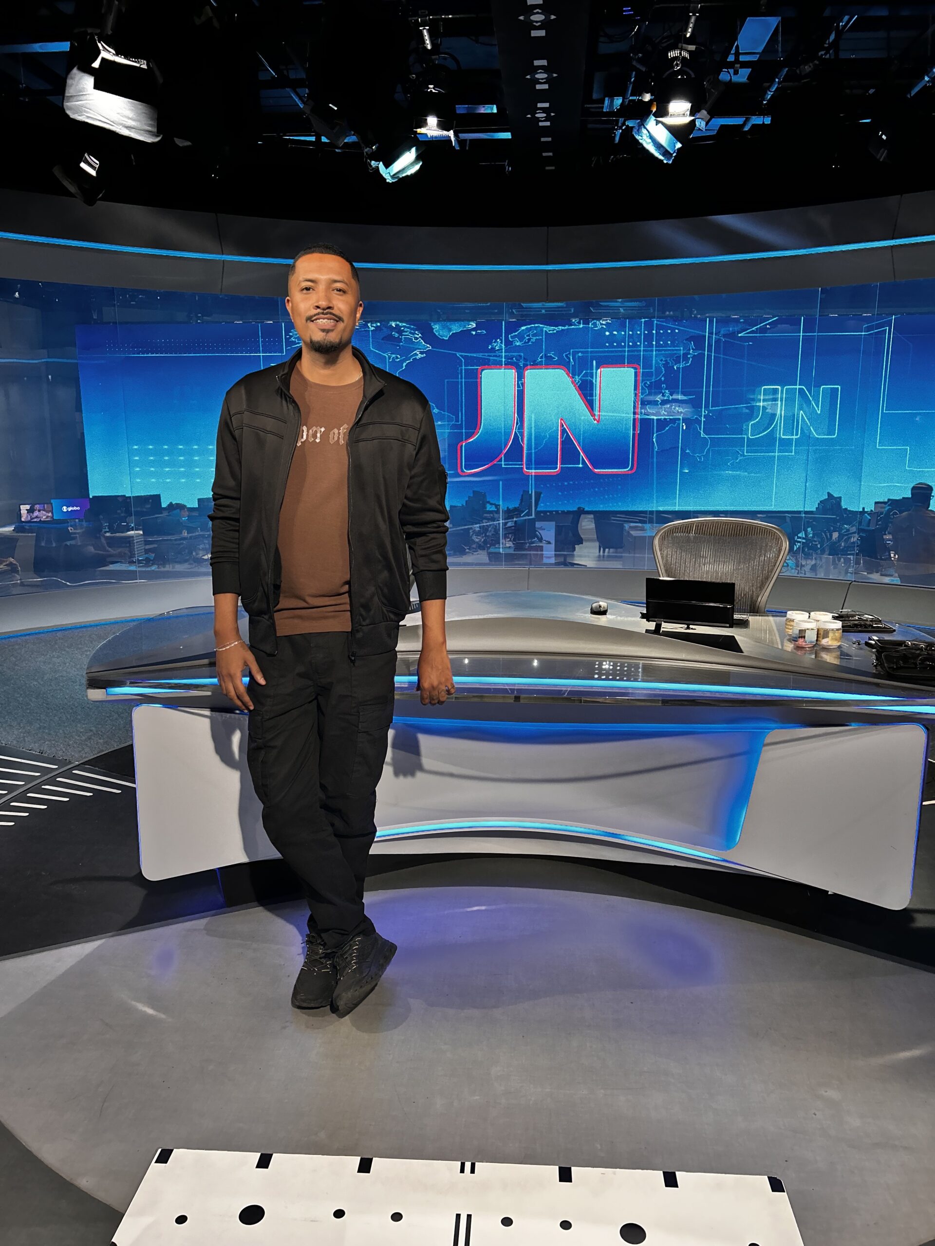 Rapaz negro vestindo um calça preta, blusa marrom e casaco preto, em pé no cenário do JN.