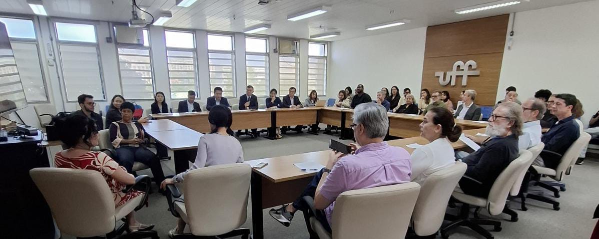 foto tirada do fundo da sala, com pessoas sentadas em uma bancada com cadeiras em forma retangular