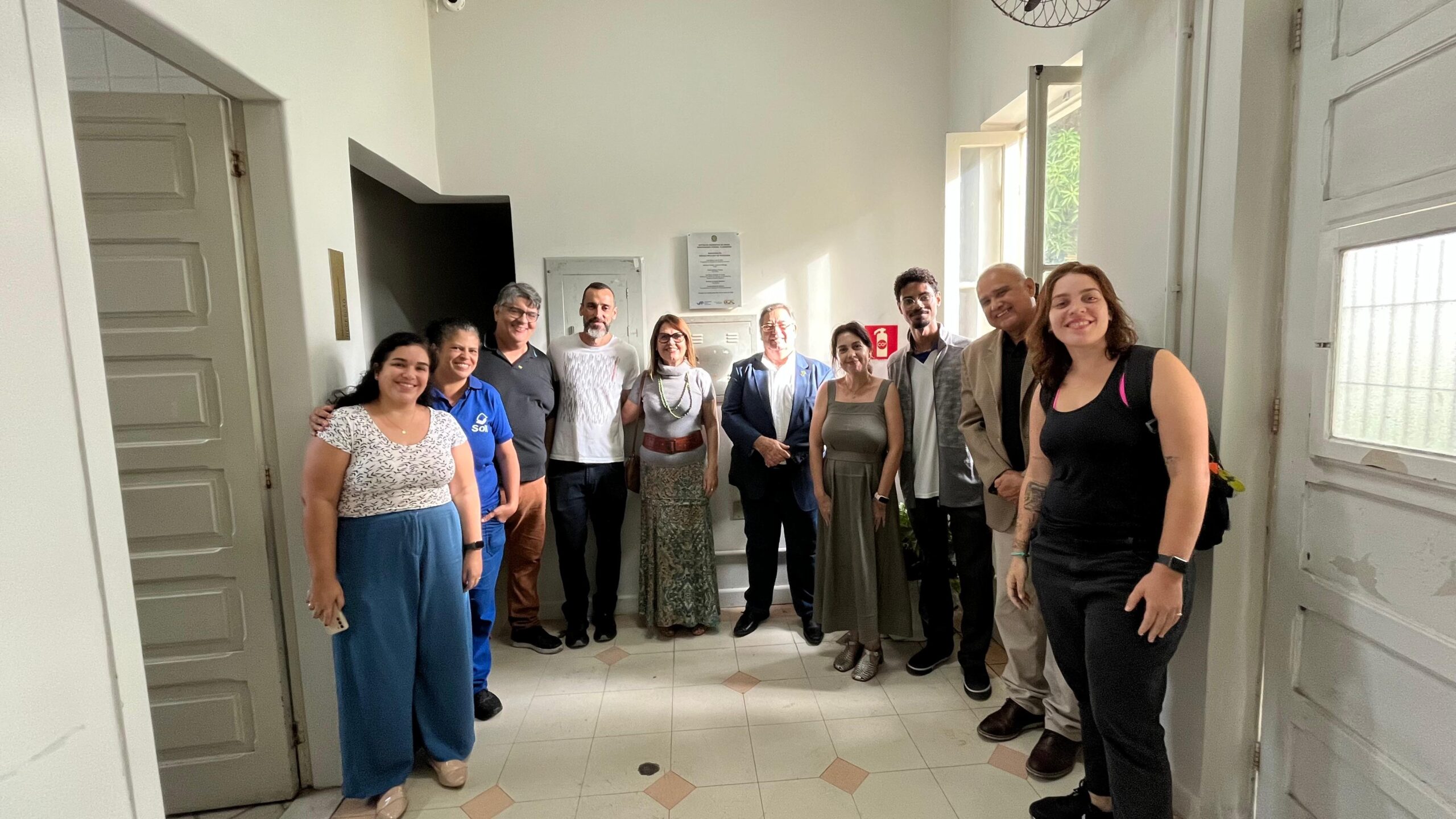 foto de um grupo de pessoas em uma sala. Ao fundo, na parede, está a placa de inaurução do espaço