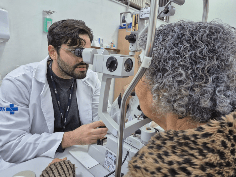 homem vestindo jaleco branco, usando um equipamento para observar os olhos de uma senhora de cabelo grisalho.