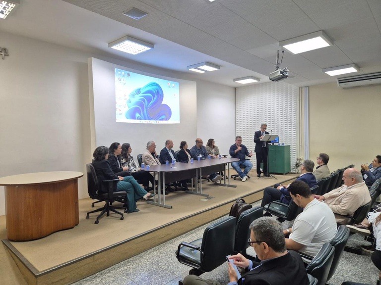foto de um auditório com pessoas na plateia, uma mesa solene e uma projeção atrás