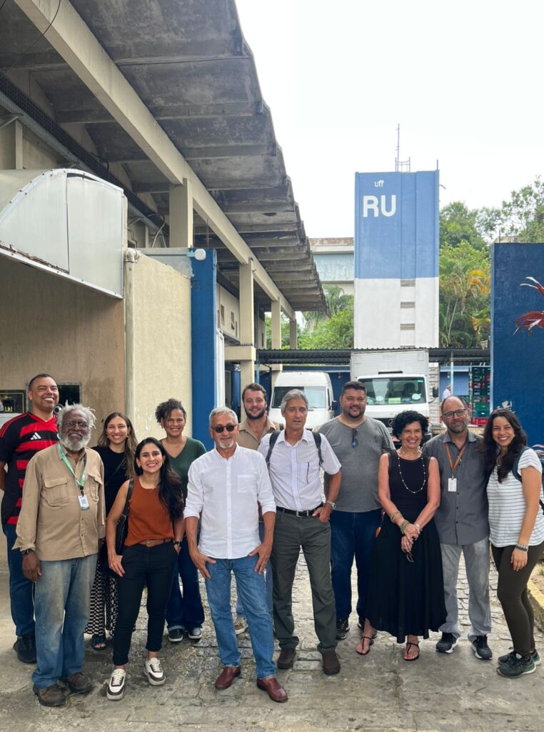 Grupo de pessoas posa em pé para foto em área externa, em frente a um prédio.Ao fundo, há um totem azul com a sigla “RU” e veículos estacionados.