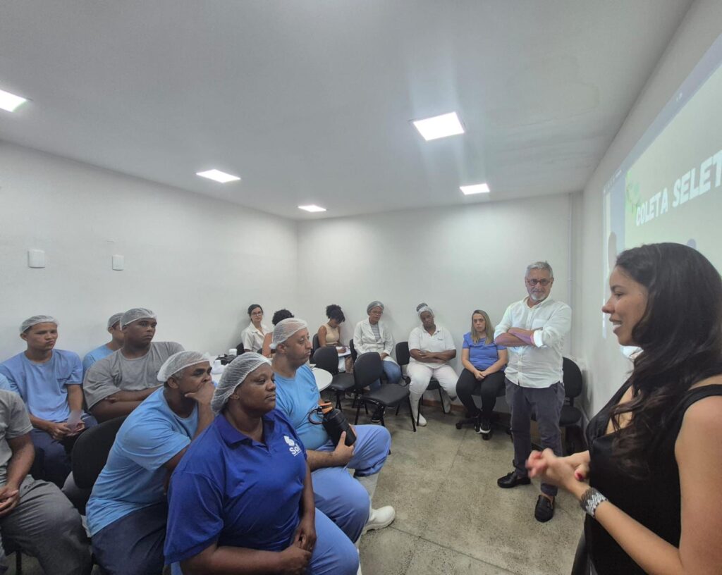 Sala clara com várias pessoas sentadas, a maioria usando uniformes azuis e toucas, atentas a uma apresentação.À direita, uma mulher em pé fala e gesticula para o grupo.
Ao fundo, há uma projeção com o texto “Coleta Seletiva”.