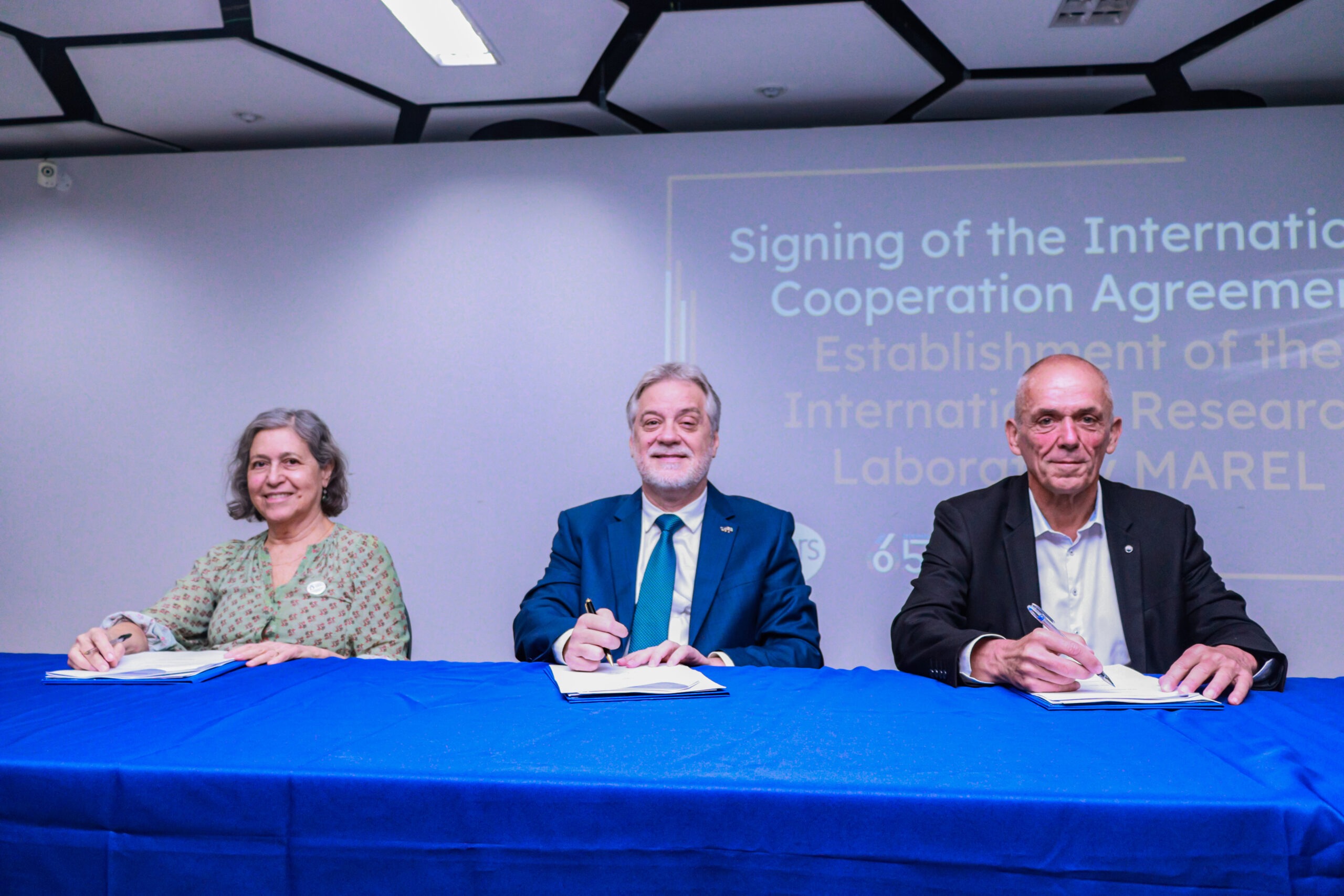 IMG_5294 Três pessoas estão sentadas atrás de uma mesa com toalha azul, assinando documentos durante uma cerimônia institucional. Ao centro, um homem de terno azul; à esquerda, uma mulher de blusa verde; à direita, um homem de terno escuro. Ao fundo, uma tela projeta texto sobre a assinatura de um acordo de cooperação internacional.
