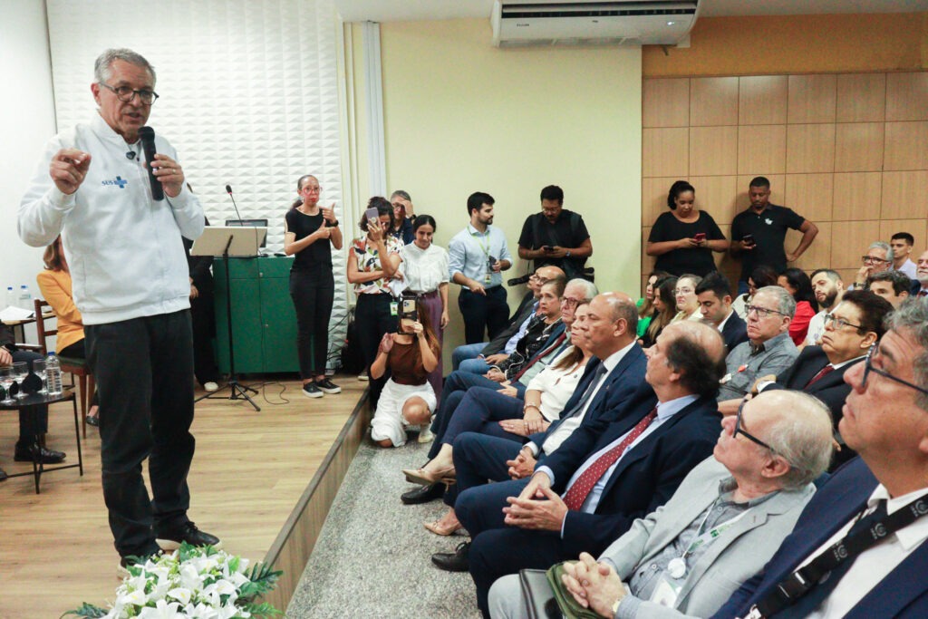 Imagem mostra um auditório lotado, com um homem em pé à frente, usando jaleco branco e falando ao microfone.O público está sentado, atento, enquanto profissionais registram o momento com celulares e câmeras.