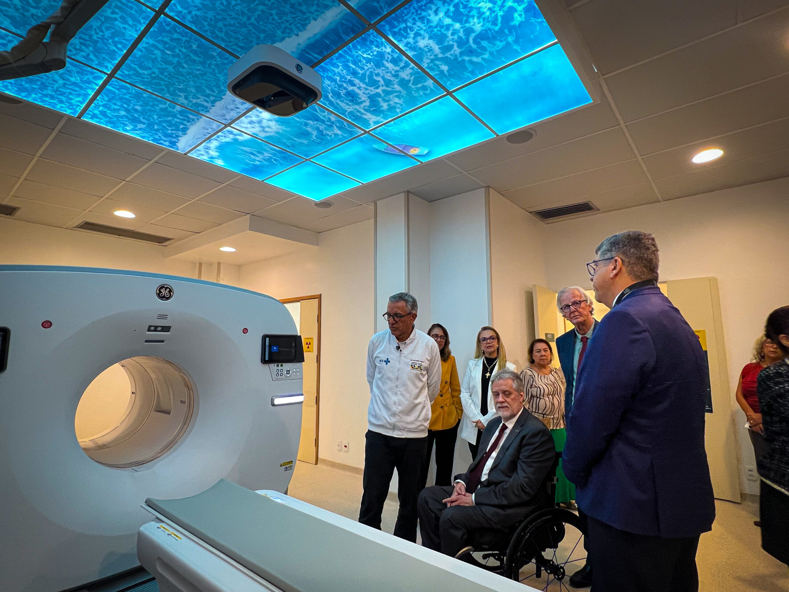 Imagem mostra uma sala hospitalar com um grande equipamento de tomografia (PET-CT) em destaque, à esquerda. À direita, um grupo de pessoas, incluindo profissionais de saúde e autoridades, observa o equipamento durante uma visita institucional.
