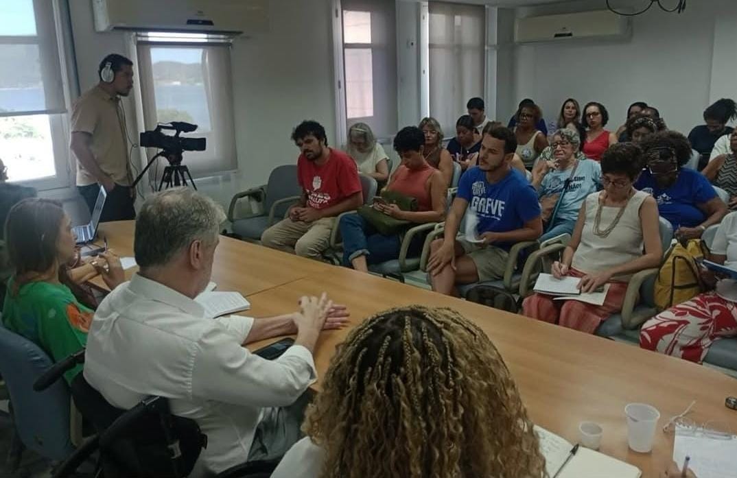 Sala de reunião com cerca de vinte pessoas sentadas em cadeiras voltadas para uma mesa retangular, onde alguns participantes conversam e fazem anotações. À esquerda, um cinegrafista grava o encontro com uma câmera em tripé, enquanto a plateia acompanha atentamente a discussão.
