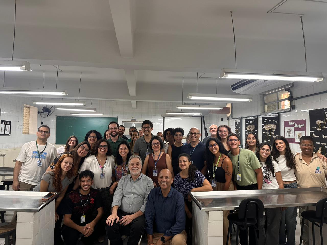 Grupo grande de pessoas reunidas em uma sala de aula ou laboratório, posando juntas e sorrindo para a foto. Ao fundo, há quadro, cartazes científicos e iluminação fluorescente no teto. Alguns estão em pé e outros agachados à frente, criando um clima de integração e registro coletivo de uma atividade acadêmica.