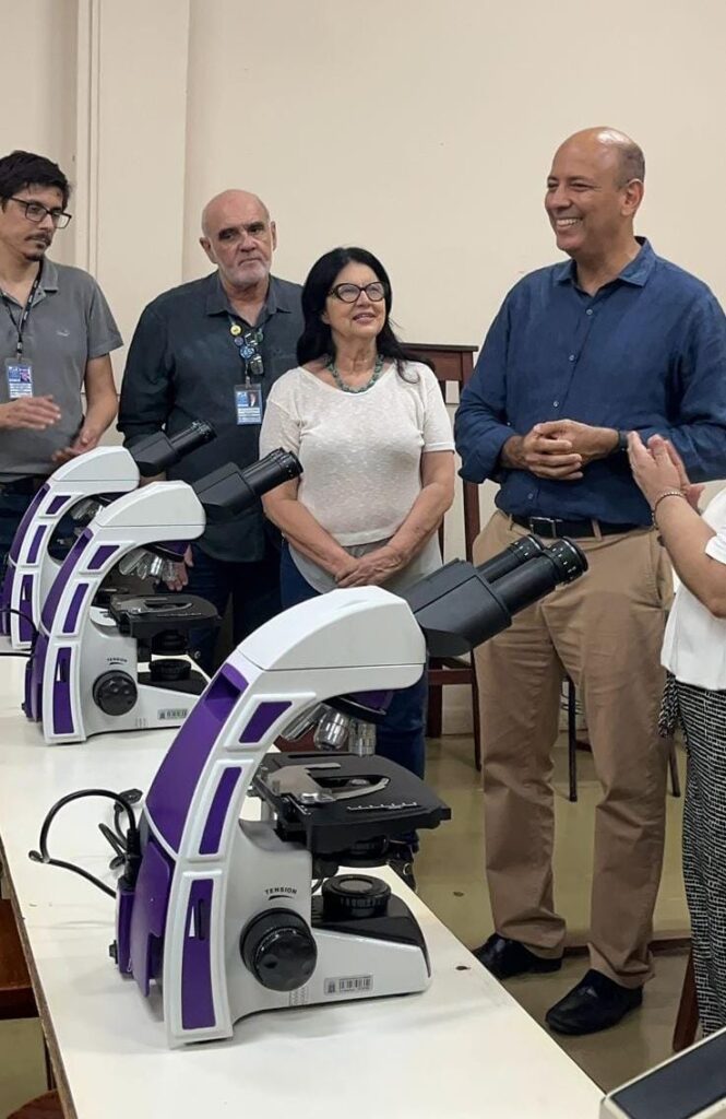 Ambiente interno de laboratório ou sala de aula, com três microscópios alinhados sobre uma mesa em primeiro plano. Ao fundo, quatro adultos em pé observam e conversam, com expressão atenta e sorridente. Os microscópios são brancos com detalhes roxos, e o grupo veste roupas casuais, sugerindo uma apresentação ou atividade prática científica.
