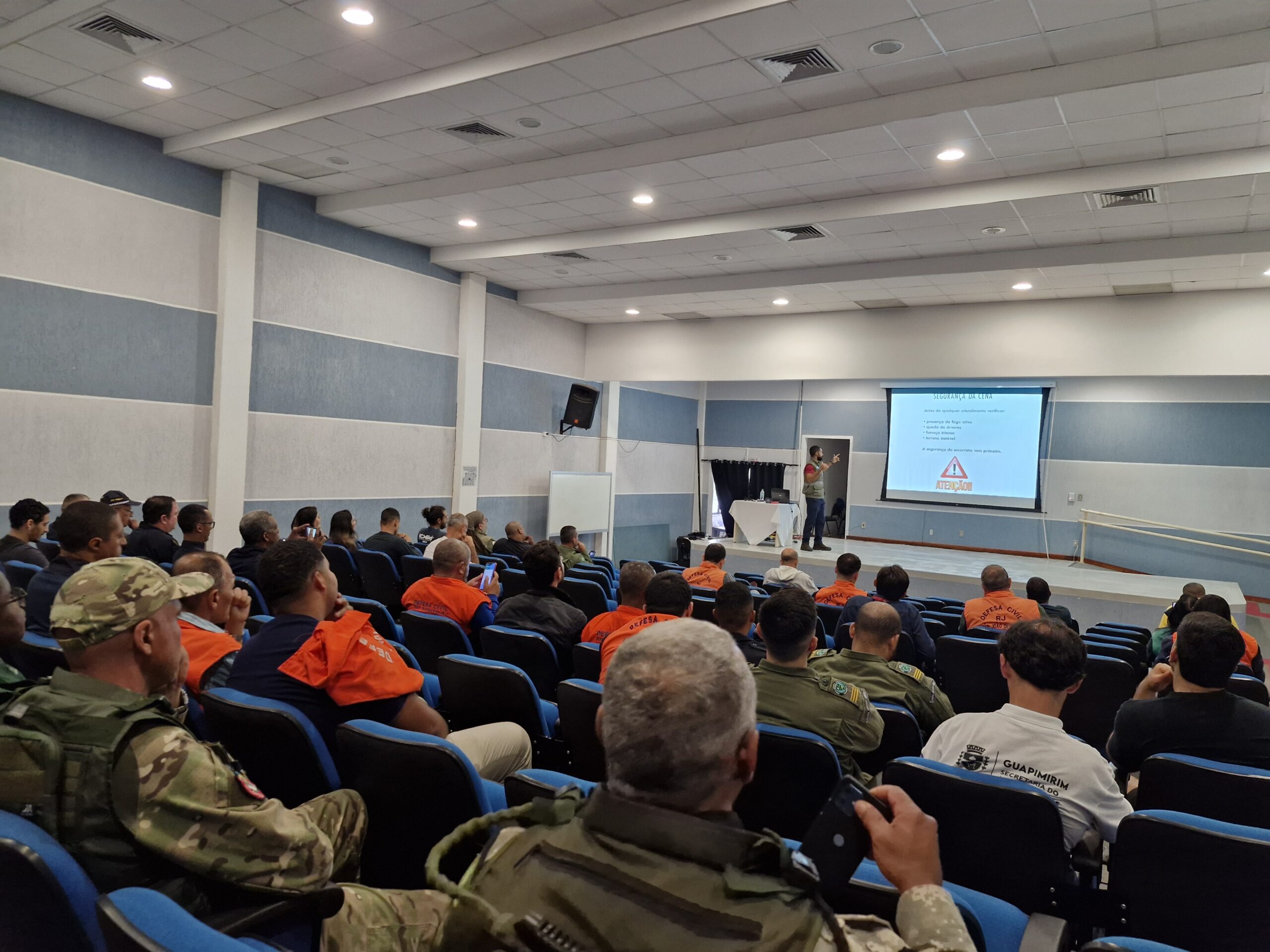 Auditório do Instituto de Física da UFF durante treinamento de prevenção e combate a incêndios florestais promovido pela UFF e pelo INEA-RJ. Na imagem, participantes entre guarda-parques, agentes da Defesa Civil e gestores ambientais acompanham uma palestra exibida em telão no palco.