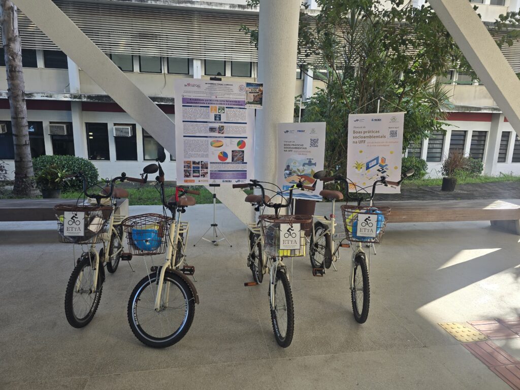 Quatro bicicletas compartilhadas do projeto UFF de Bike estão estacionadas em uma área coberta do campus, alinhadas em frente a painéis informativos sobre sustentabilidade e boas práticas socioambientais. As bicicletas são claras, com cestos frontais e placas de identificação “ETTA”. Ao fundo, aparecem bancos de madeira, jardins e o prédio da universidade com janelas e aparelhos de ar-condicionado. A iluminação natural destaca o ambiente amplo e arborizado.