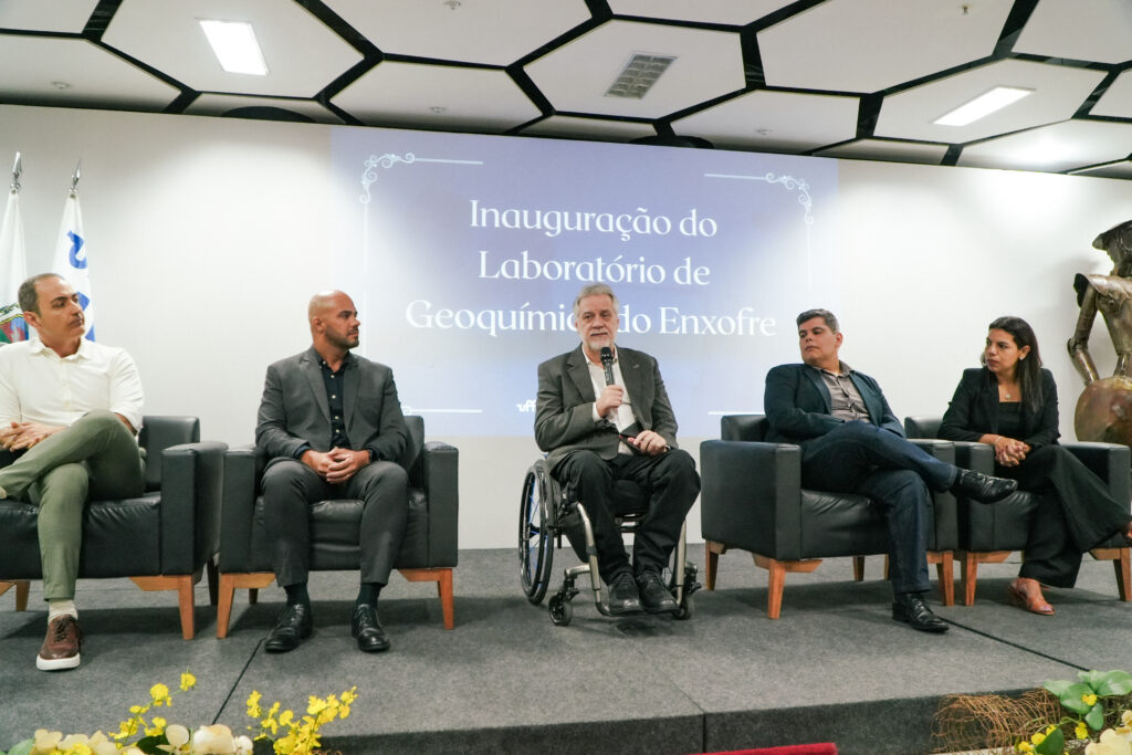 Mesa com cinco participantes em evento de inauguração, com um homem ao centro falando ao microfone diante de projeção ao fundo.
