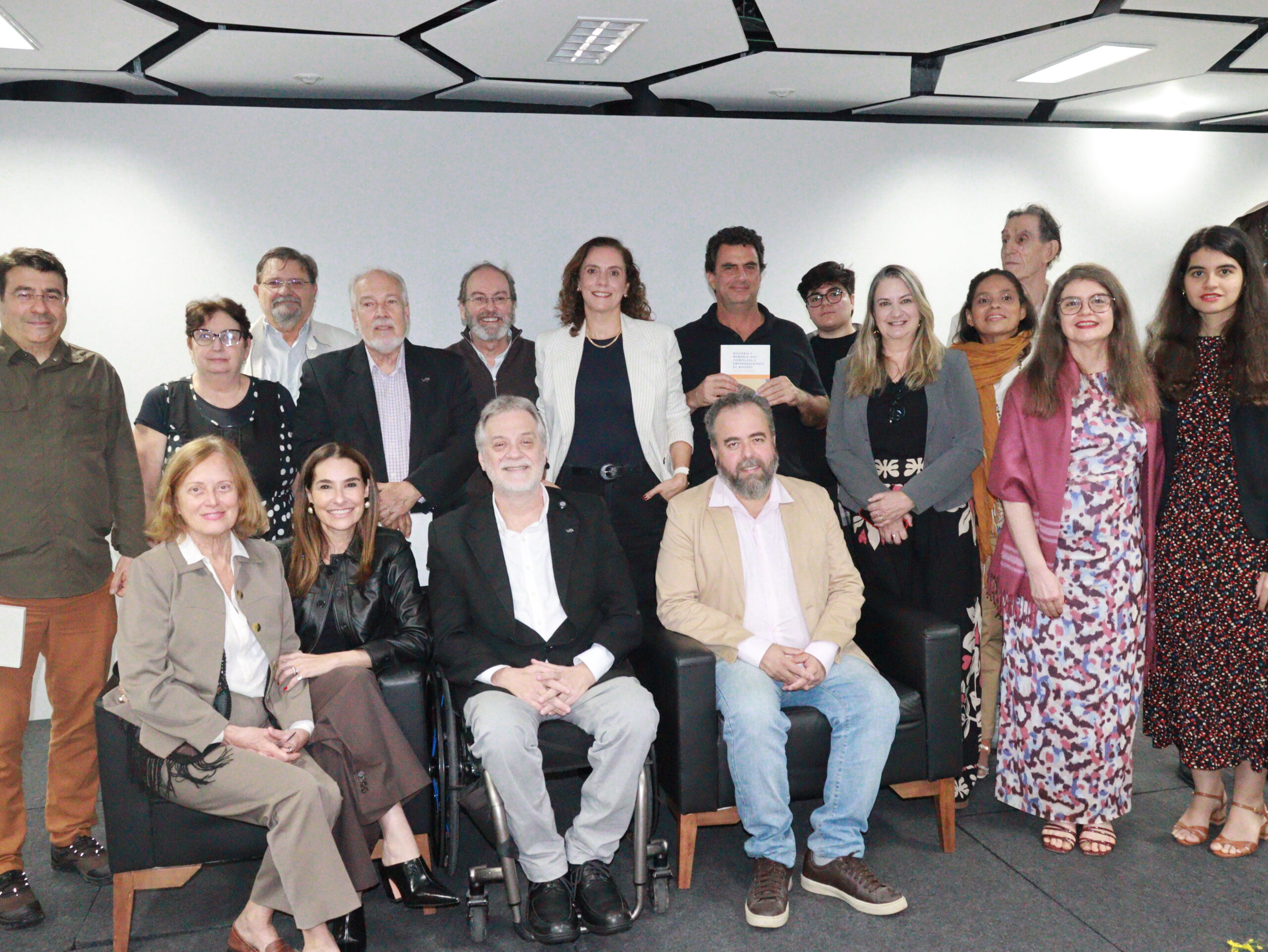 Foto de grupo em ambiente interno, com cerca de quinze pessoas de diferentes idades posando juntas diante de uma parede branca. Na frente, três pessoas estão sentadas — uma delas em cadeira de rodas — e as demais aparecem em pé atrás, sorrindo para a câmera. O grupo usa roupas sociais e casuais em tons variados. Ao centro, uma mulher de blazer claro e um homem segurando um livro se destacam. O espaço tem iluminação forte e teto moderno com painéis geométricos brancos e pretos.