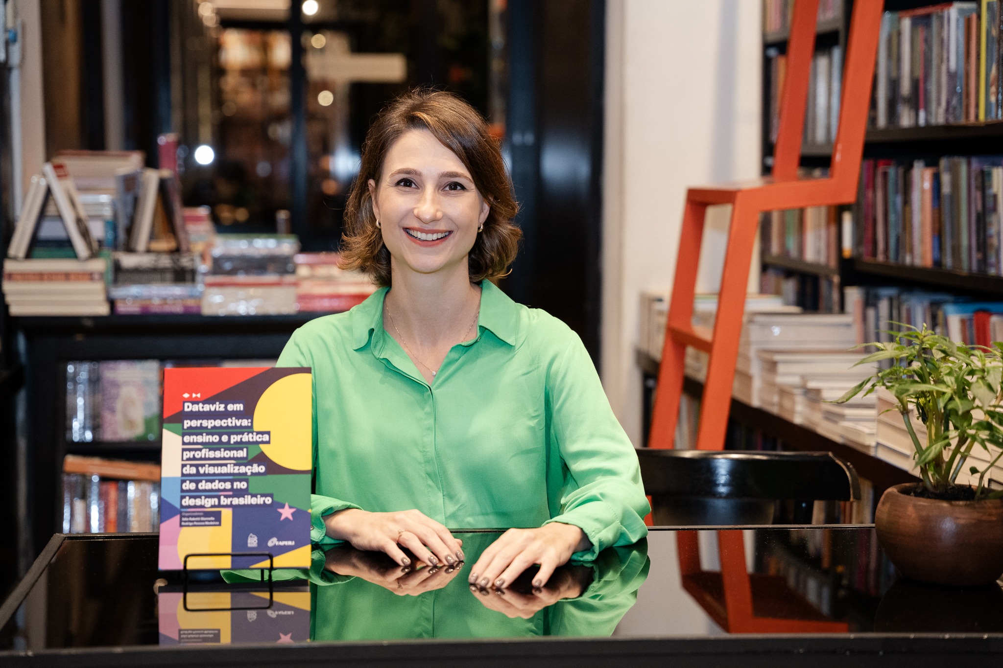 Lançamento do Livro: Dataviz em Perspectiva: ensino e prática Mulher branca, de cabelos curtos castanhos, sentada à mesa em uma livraria ou biblioteca, sorrindo para a câmera. Ela veste camisa verde-clara e está ao lado do livro Dataviz em perspectiva: ensino e prática profissional da visualização de dados no design brasileiro, apoiado sobre a mesa. Ao fundo, há estantes repletas de livros e uma escada vermelha decorativa.
