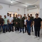 Grupo de aproximadamente 14 pessoas posa em sala ampla e iluminada, algumas usando jalecos verdes, em ambiente de formação técnica.