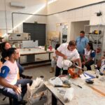 Grupo de profissionais acompanha demonstração técnica em oficina equipada com ferramentas, máquinas e materiais de trabalho.