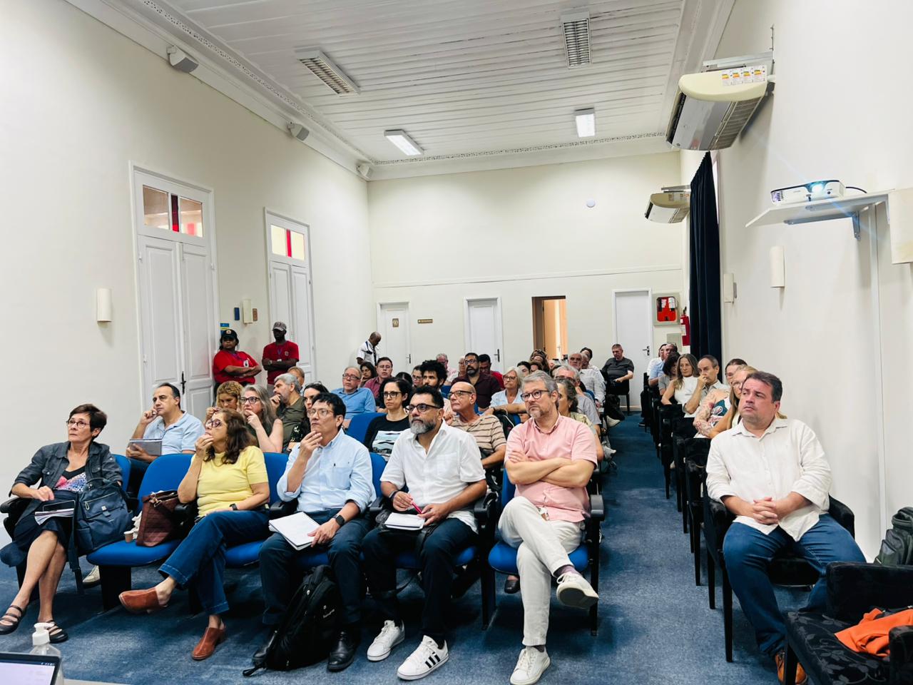 Fotografia mostra uma sala de reuniões da UFF com dezenas de participantes sentados em cadeiras azuis, acompanhando atentamente uma apresentação fora do enquadramento. O ambiente é amplo, com paredes claras, teto alto e iluminação branca. Na primeira fila, homens e mulheres aparecem com notebooks, cadernos e mochilas, em postura de escuta e concentração. Ao fundo, outras pessoas acompanham o encontro, reforçando o caráter institucional e coletivo da reunião.