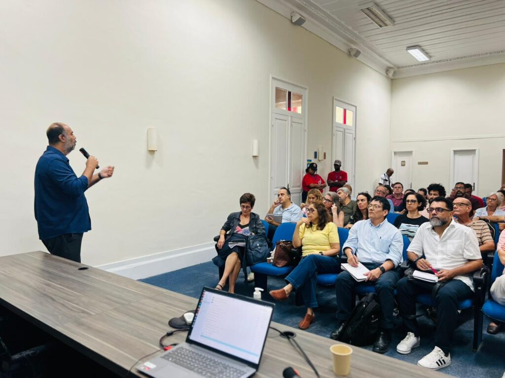 otografia mostra um homem em pé, à esquerda da imagem, falando ao microfone durante uma reunião da UFF. À frente dele, dezenas de participantes estão sentados em cadeiras azuis, atentos à apresentação. Sobre a mesa em primeiro plano há um notebook aberto e materiais de apoio. O ambiente é amplo, com paredes claras, teto alto e iluminação branca, reforçando o caráter institucional do encontro.