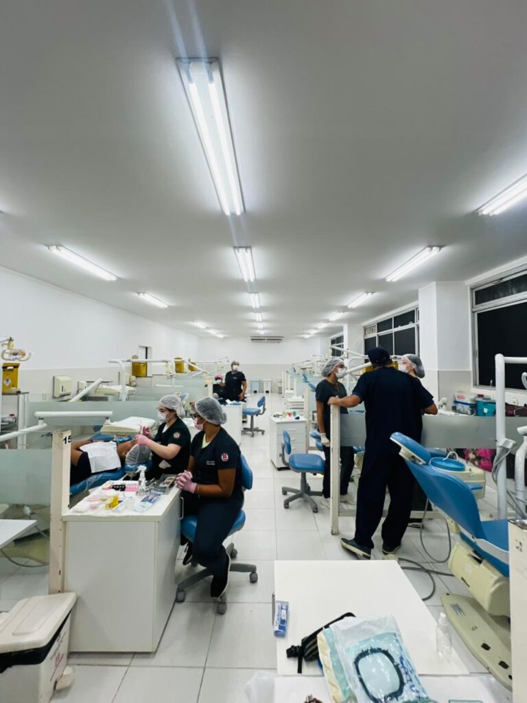 fotografia em ambiente interno de clínica odontológica coletiva. A sala é ampla, comprida e iluminada por várias luminárias fluorescentes instaladas no teto branco. Nas laterais e ao centro do espaço há cadeiras odontológicas em tons de azul-claro e branco, acompanhadas de equipamentos clínicos.