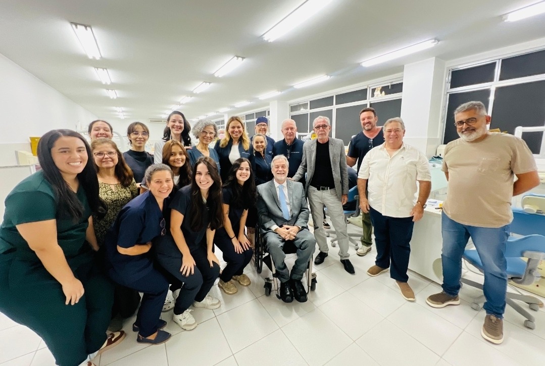 fotografia em uma clínica odontológica coletiva, com iluminação branca intensa e cadeiras odontológicas ao fundo. Ao centro da imagem, um grupo de cerca de quinze pessoas posa para a foto, entre estudantes, profissionais e convidados. A maioria veste roupas escuras ou jalecos, enquanto alguns homens usam roupas sociais ou casuais claras. Todos sorriem para a câmera, em um ambiente que transmite integração acadêmica e celebração coletiva.