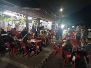 Área externa de um bar à noite, com público distribuído em mesas vermelhas na calçada acompanhando uma palestra do Pint of Science. Ao fundo, uma pessoa fala ao microfone em frente ao estabelecimento iluminado, enquanto participantes assistem ao debate em clima informal.