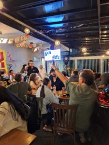 Ambiente interno de bar com várias pessoas sentadas em mesas de madeira durante uma atividade do Pint of Science. Ao centro, um participante levanta o braço enquanto acompanha a conversa. O espaço tem iluminação amarelada, decoração descontraída e uma televisão exibindo imagens ao fundo.
