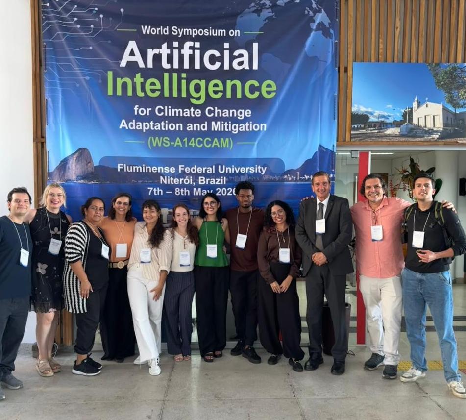 Grupo de onze pessoas posa em pé em um espaço interno da UFF, em frente a um grande painel azul do simpósio internacional “World Symposium on Artificial Intelligence for Climate Change Adaptation and Mitigation”, realizado em Niterói. Os participantes usam crachás de identificação e sorriem para a foto. Ao fundo, além do banner com elementos gráficos de tecnologia e imagem do Pão de Açúcar, há divisórias de madeira e uma fotografia emoldurada na parede.