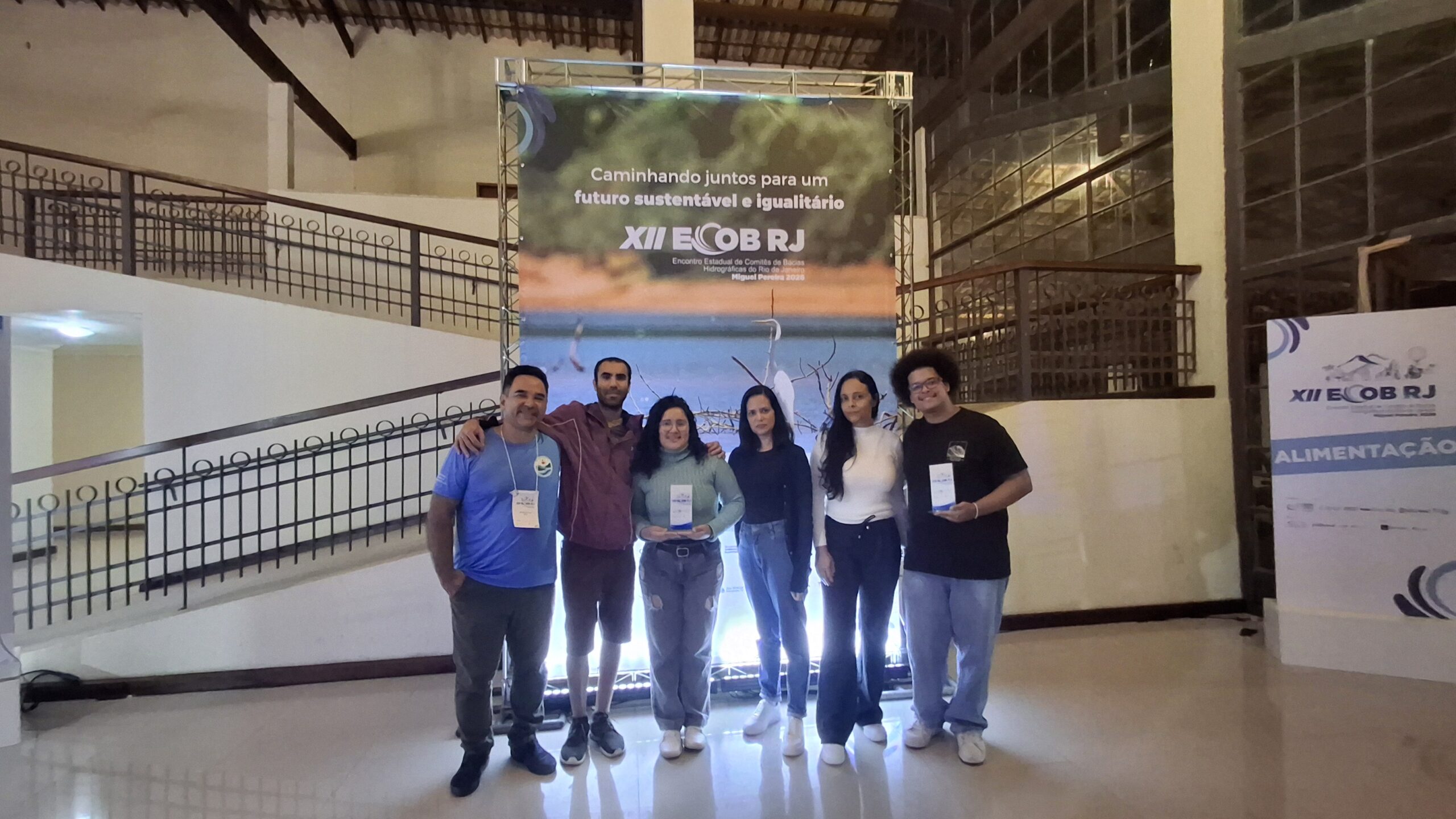 20260415_182212 Na imagem, seis pessoas posam lado a lado em um espaço interno amplo de um centro de eventos, em frente a um painel iluminado do XII ECOB-RJ (Encontro Estadual de Comitês de Bacias Hidrográficas do Rio de Janeiro), que exibe o slogan “Caminhando juntos para um futuro sustentável e igualitário”. Duas integrantes do grupo seguram troféus ou placas de premiação. Ao fundo, observam-se uma estrutura metálica de sustentação do painel, mezaninos com guarda-corpos, grandes janelas envidraçadas e teto com vigas aparentes de madeira. À direita da cena, há um totem com a identidade visual do evento. O ambiente é bem iluminado e transmite o contexto de uma cerimônia ou registro institucional após a premiação.
